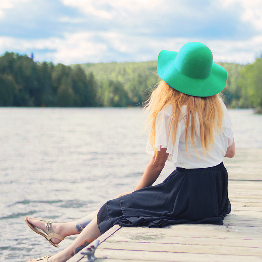 Woman with POTS sitting on a lake dock wearing a turquoise hat and drinking NormaLyte
