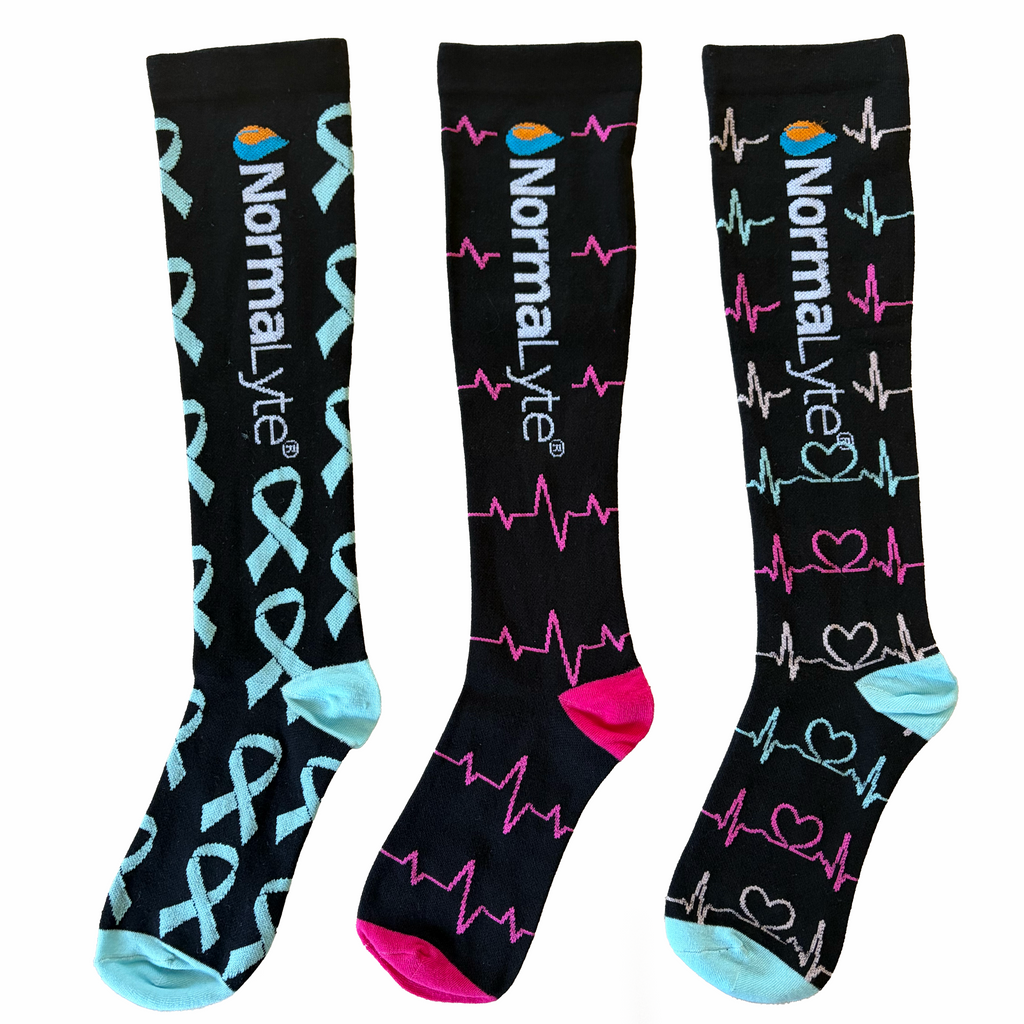 Three black NormaLyte graduated compression socks displaying medical-themed patterns, including teal awareness ribbons, pink EKG heartbeat lines, and a mix of heart and heartbeat designs