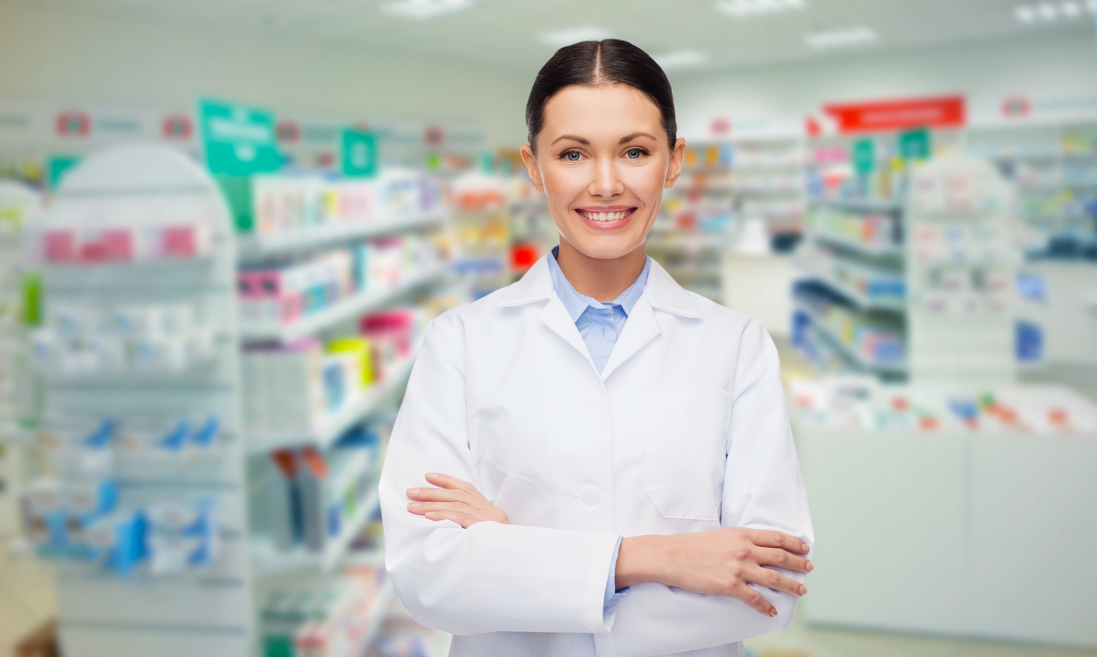 Smiling female pharmacist in a white lab coat stands with arms crossed inside a modern pharmacy.
