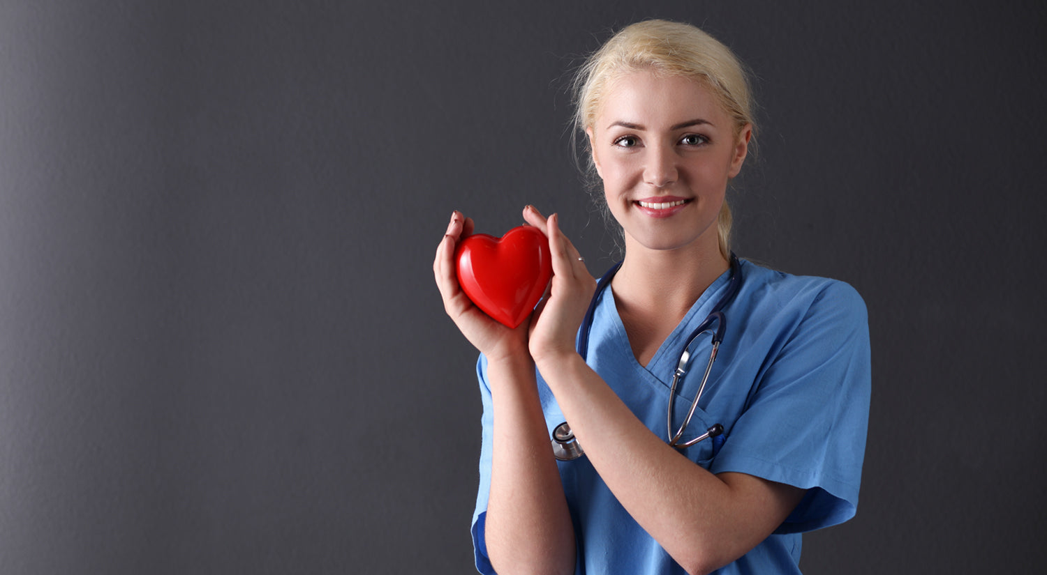 Smiling nurse in blue scrubs with a stethoscope holds a red heart against a dark gray background. 