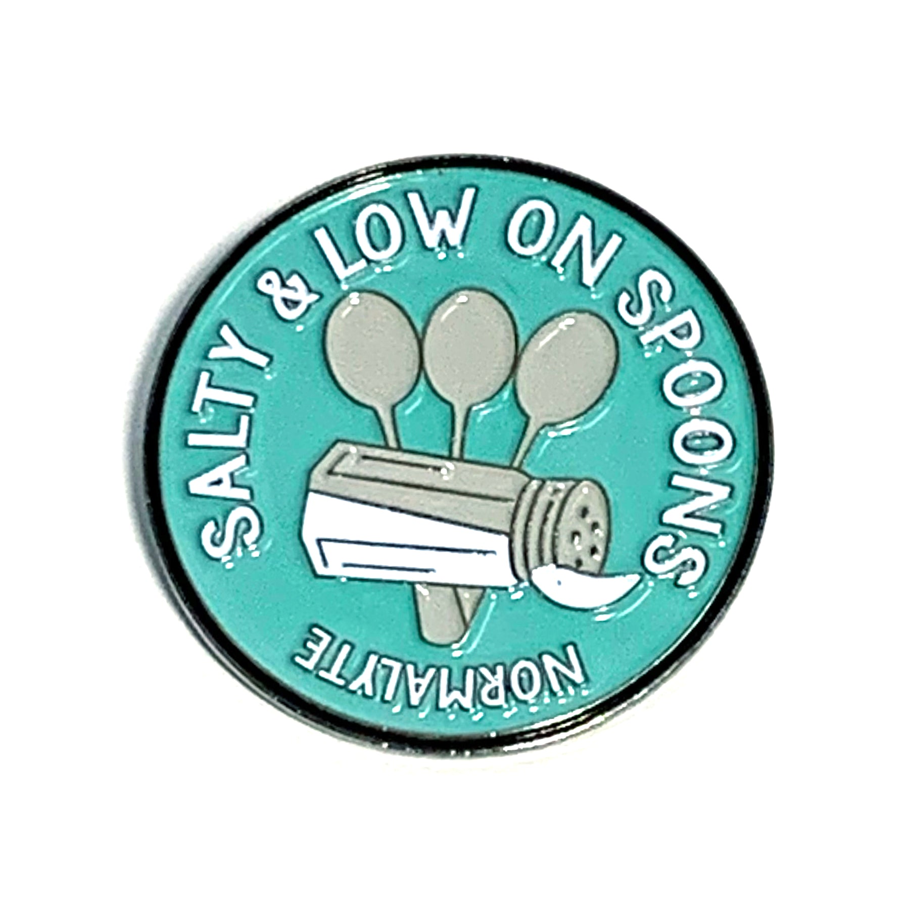 Close-up of the round teal NormaLyte enamel lapel pin showing the detailed "Salty & Low on Spoons" design featuring a salt shaker and three spoons